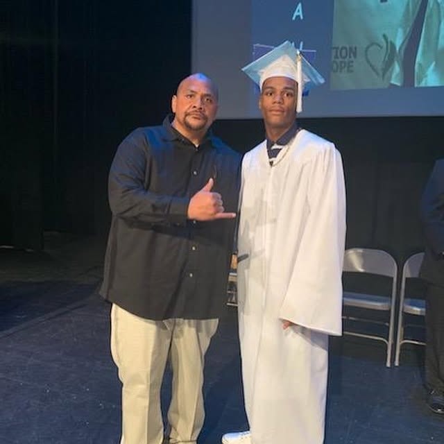 Coach and Class of 2019 graduate in white cap and gown posing on stage at Operation New Hope ceremony