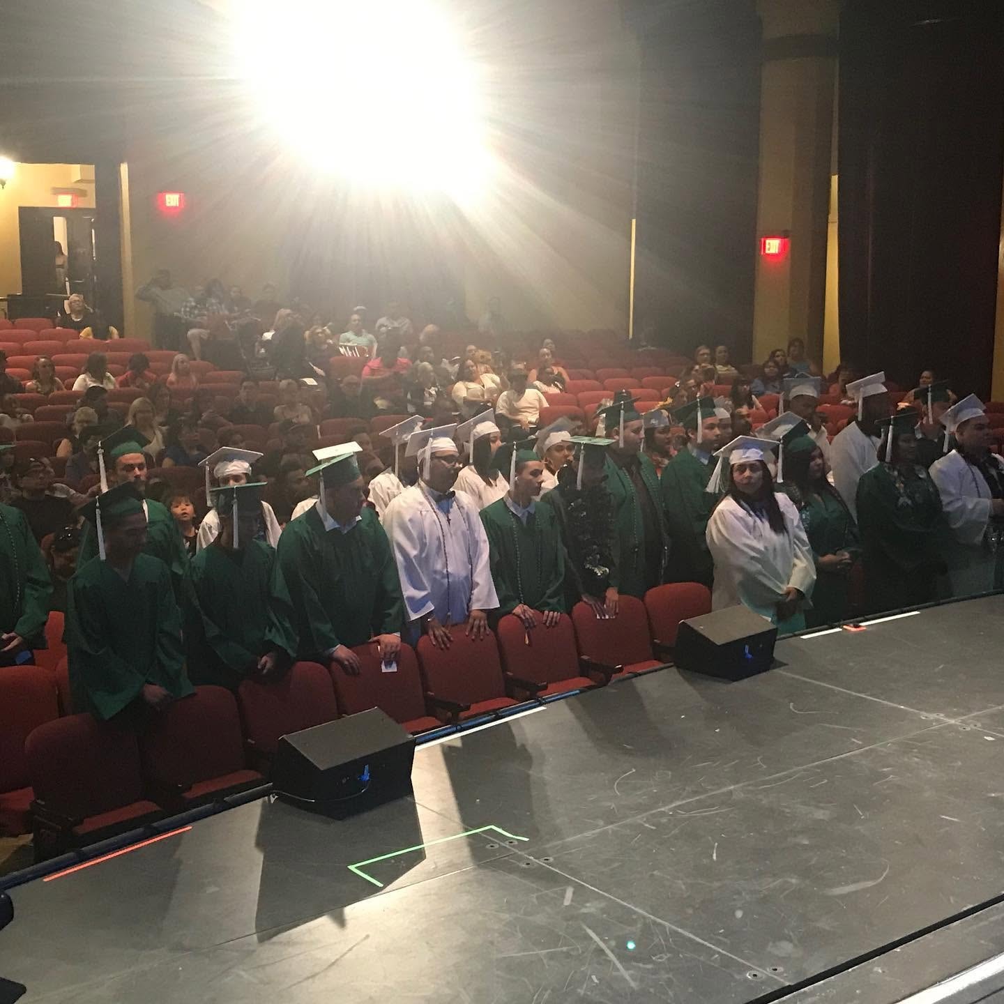 Class of 2019 graduates in green and white caps and gowns standing in packed auditorium at ceremony