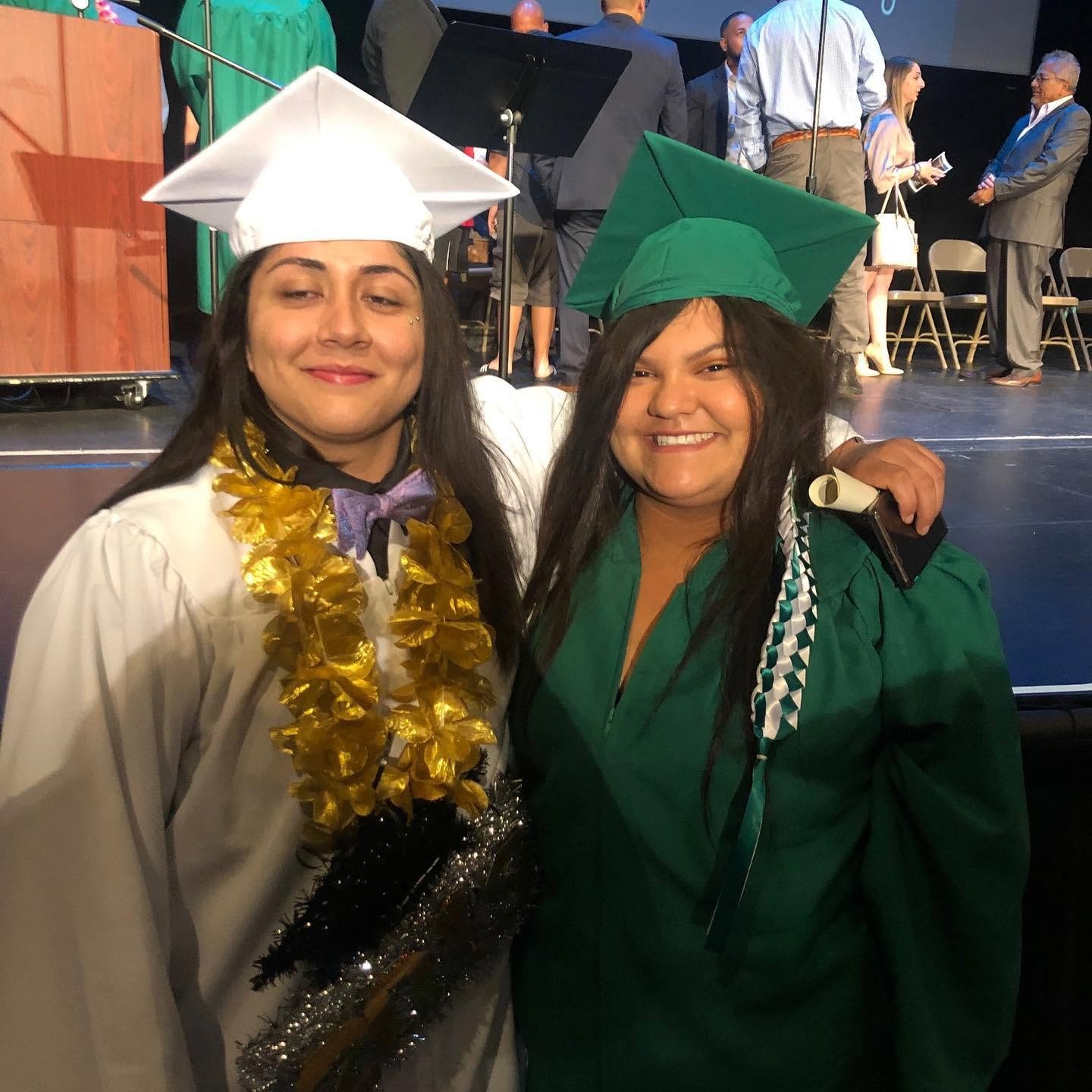 Two Class of 2019 graduates in white and green caps and gowns smiling at ceremony